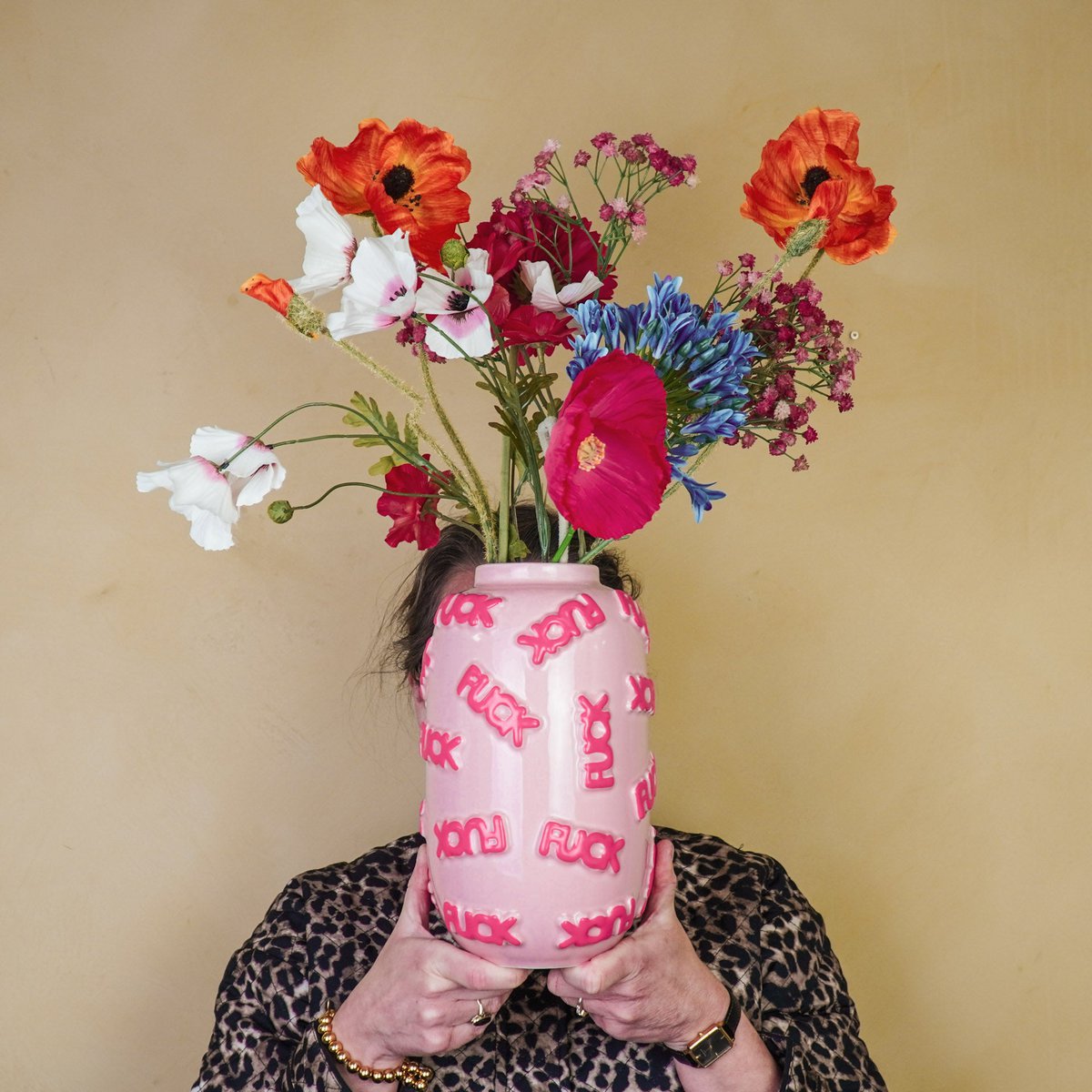 Persoon die de roze Fuck vaas met kleurrijke bloemen voor het gezicht houdt tegen een sfeervolle geel-bruine muur.