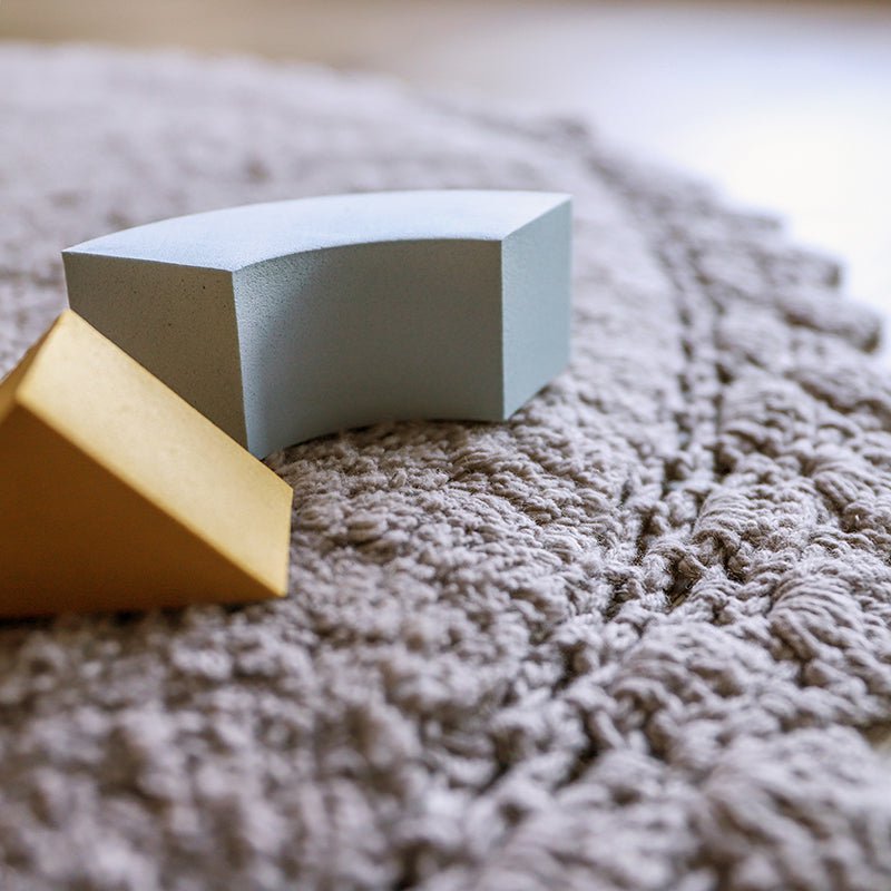 Detailopname van het zachte, grijze oppervlak van het handgetufte Perla vloerkleed met twee houten bouwblokjes erop.
