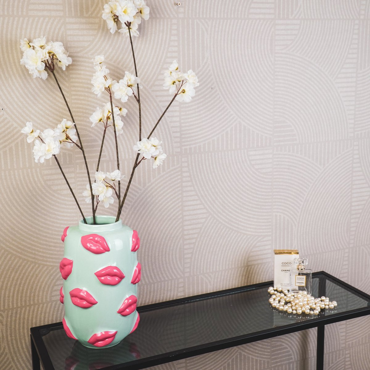 De mintgroene lipvaas met witte bloemen op een glazen tafel tegen een warme beige achtergrond.