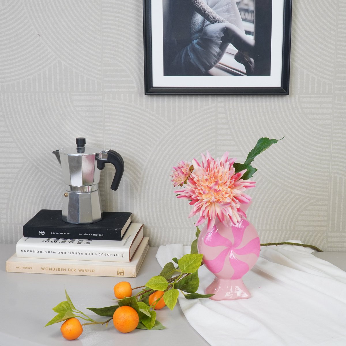 Roze Toffee Candy vaas met bloemen op een bureau naast een tak met mandarijnen tegen een beige muur.