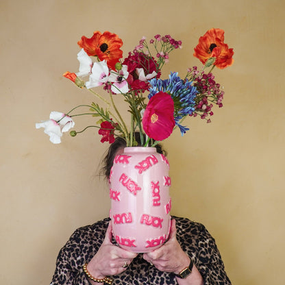 Persoon die de roze Fuck vaas met kleurrijke bloemen voor het gezicht houdt tegen een sfeervolle geel-bruine muur.